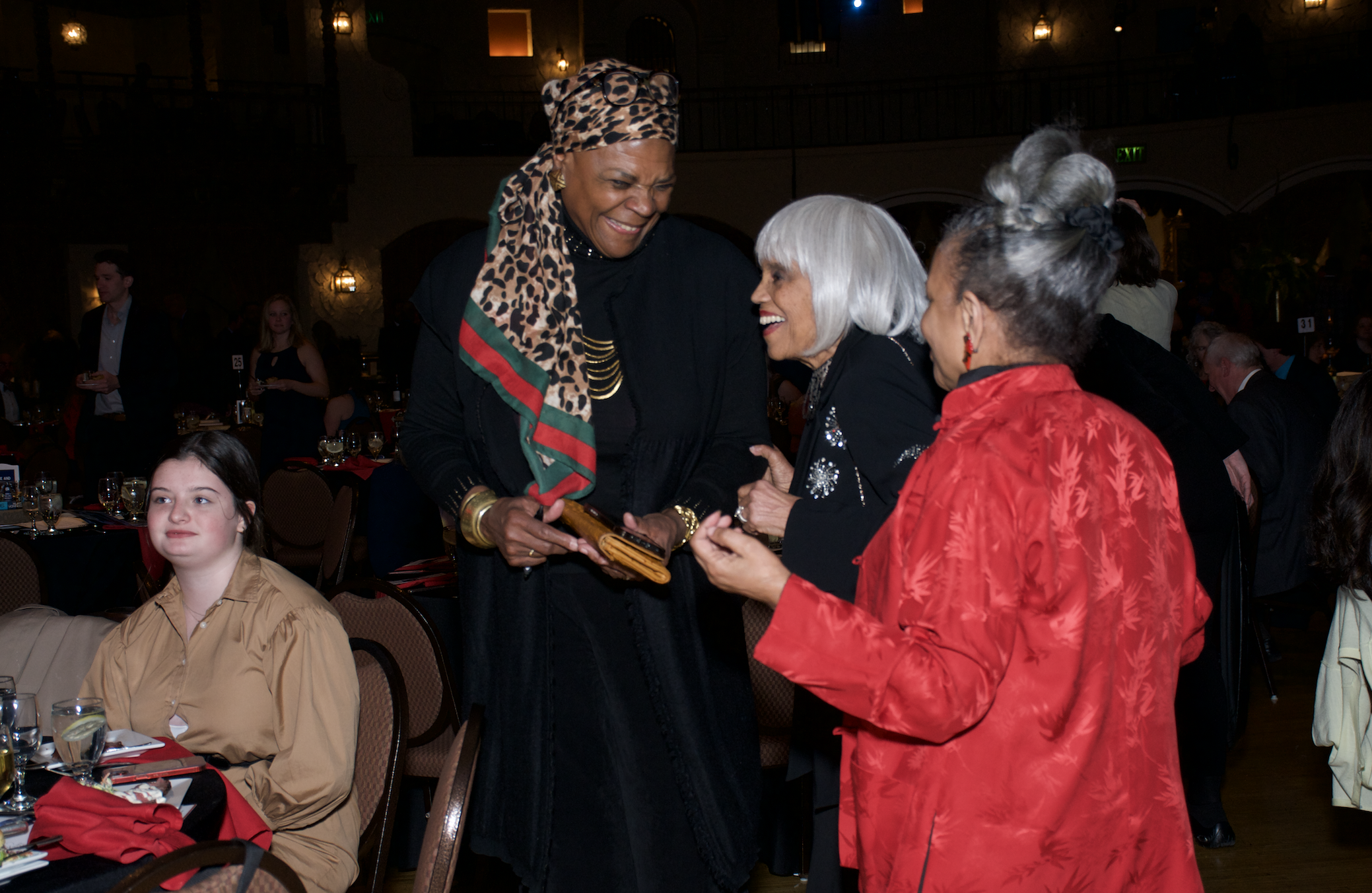 Three people talking together at a gathering of many people